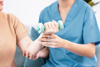 Caregiver helping contented senior woman exercise with dumbbell at home. Rehab for elder, senior medical care.