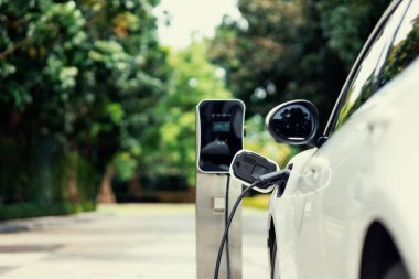 Focus closeup electric vehicle plugged in with EV charger device from blurred background of public charging station powered by renewable clean energy for progressive eco-friendly car concept.