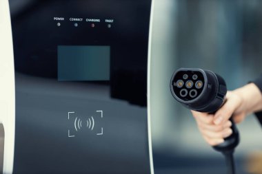 Focus closeup hand holding EV plug for electric vehicle pointing at camera with charging station at background as progressive idea of alternative sustainable clean energy for ecological awareness idea