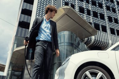 Below view of progressive businessman with electric car recharging at public charging station at modern city residential building. Eco friendly rechargeable EV car powered by alternative clean energy.