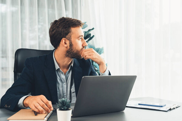 Businessman in black suit working on laptop at his workspace desk. Smart executive researching financial data and planning marketing strategy on corporate laptop at modern workplace. Entity