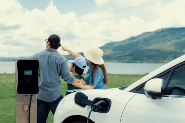 Concept of progressive happy family enjoying their time at green field and lake with electric vehicle. Electric vehicle driven by clean renewable from eco-friendly power sauce.