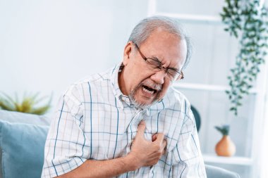 An agonizing senior man suffering from chest pain or heart attack alone in his living room. Serious health problem and feeling unwell concept for seniors.