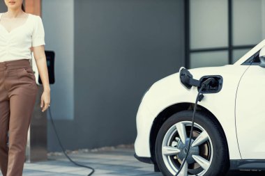 Focus image of electric vehicle recharging battery at home charging station with blurred woman walking in the background. Progressive concept of green energy technology applied in daily lifestyle.