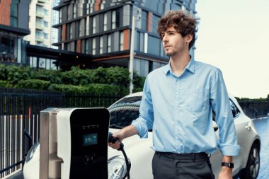 Progressive businessman insert charger plug from charging station to his electric vehicle with apartment condo building in background. Eco friendly rechargeable car powered by sustainable energy.