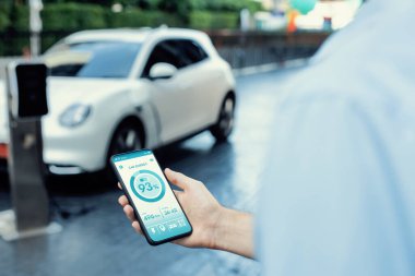Closeup progressive businessman look at EV cars battery status application on smart phone screen at public parking charging station with power cable plug and renewable energy-powered electric vehicle