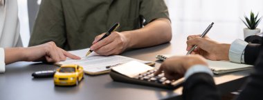 Araba satıcısı faiz oranını ve araba kredisinin maliyetlerini hesap makinesiyle hesaplayarak, satış bürosundaki anlaşma ve dönem detaylarını müşteriye açıklar ve finans ve sigorta hizmeti sunar. Dahi.