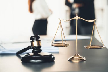 Adalet ve tokmak çekicinin dengeli ölçeğinde, avukat meslektaşlarının hukuki temsilciler olarak hukuk bürosunda görüşüp dava açmayı planladıkları bulanık arka planı üzerinde duruluyor. Eşitlik