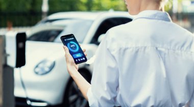 Suit-clad progressive businesswoman look at EV cars battery status from her phone while standing on a charging station with a power cable plug and a renewable energy-powered electric vehicle.