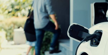 Arka planda bulanık baba ve kızının yürüdüğü şarj istasyonunda elektrikli araba şarj etmeye odaklan. Sağlıklı çevre yaşam tarzı konsepti için yenilikçi yeşil ve temiz enerji aracı.
