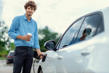 Progressive eco-friendly concept of parking EV car at public electric-powered charging station in city with blur background of businessman leaning on recharging-electric vehicle with coffee.