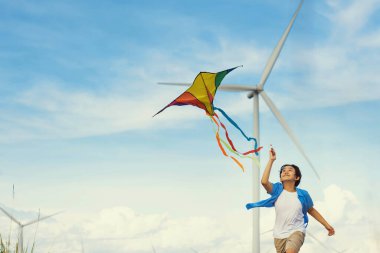 İlerici mutlu kaygısız çocuk konsepti. Yol boyunca koşan ve uçurtma uçuran genç bir çocuk kırsal alanda doğal manzaralı, açık gökyüzü ve güneşli bir gün dağ ve rüzgar türbini arka planında