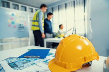 Concept of engineering equipments of protective helmet for competent engineer on piles of paper works. Blurred image of engineers and employer working together at the desk.