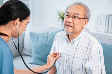 Caring young female doctor examining her contented senior patient with stethoscope in living room. Medical service for elderly, elderly sickness, declining health.