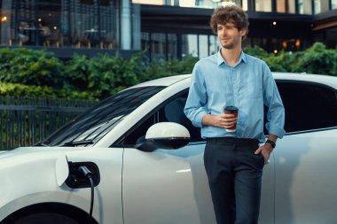 Progressive eco-friendly concept of parking EV car at public electric-powered charging station in city with blur background of businessman leaning on recharging-electric vehicle with coffee.