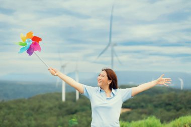 Concept of progressive happy woman relaxing at the wind turbine farm with windmill toy in her hand. Electric generator from wind by wind turbine generator on the country side with mountain on horizon.