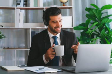 Çağrı merkezi operatörünün müdürü bilgisayarla uğraşırken masasında kahvesiyle oturuyor. Kulaklık takan ve siyah takım giyen bir ofis çalışanı müşteri desteği ya da tele pazarlama üzerinde çalışıyor. coşkulu