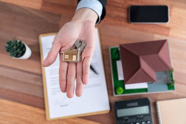 Gayrimenkul acentesinin üst düzey anahtarı, ev kredisi veya ipoteği sembolik olarak hesap makinesi ve yasal belge ile çevrili ahşap masayı ev satışları için ikna edici bir jest yaparak teslim ediyor. Sevinçli.