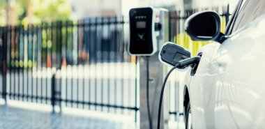 Focus closeup electric vehicle plugged in with EV charger device from blurred background of public charging station powered by renewable clean energy for progressive eco-friendly car concept.