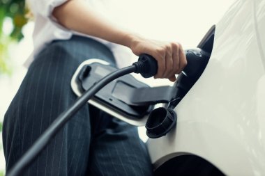 Focus EV charger plug and electric car at public charging station with blur progressive businesswoman holding charger and apartment condo building in background. Eco friendly electric vehicle concept.