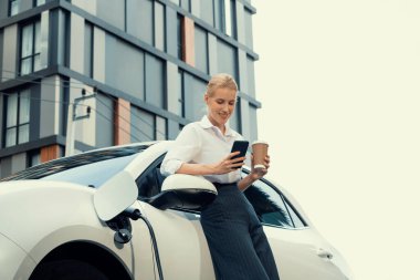 Focus businessman using phone, leaning on electric vehicle, holding coffee with blurred city residential condo buildings in background as progressive lifestyle by renewable and sustainable EV car.