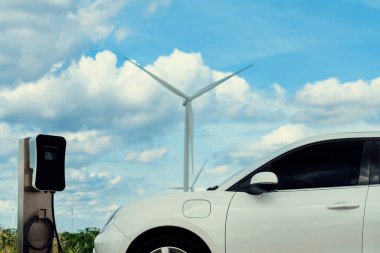 Progressive combination of wind turbine and EV car, future energy infrastructure. Electric vehicle being charged at charging station powered by renewable energy from wind turbine in the countryside.