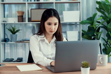 Modern ofis masasındaki Asyalı hevesli genç iş kadını bir fincan kahveyle çalışmak için dizüstü bilgisayar kullanıyor. Çalışkan ve çekici ofis hanımı ofisinde bilgisayar defteri üzerinde çalışıyor..