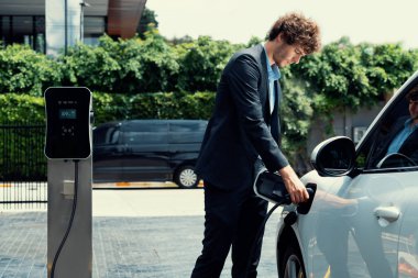Modern şehir yerleşim bölgesindeki kamu şarj istasyonunda elektrikli araba şarj eden yenilikçi bir işadamı. Çevre dostu şarj edilebilir araba alternatif temiz enerjiyle çalışıyor..