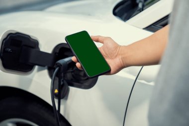 Taklit uzay için şarj istasyonuna bağlı elektrikli aracın yeşil ekran enerji durumunu gösteren bir model telefon. Temiz çevre için ilerici bir kavram. EV, yeşil yenilenebilir enerjiden güç alıyor.