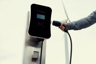 Progressive natural scenic with windmill generator where hand insert charging plug to electric vehicle from charging station with natural background. EV car powered by wind turbine electric generator.