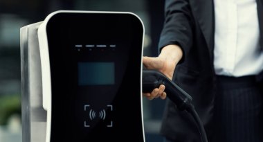 Focus EV charger plug and electric car at public charging station with blur progressive businesswoman holding charger and apartment condo building in background. Eco friendly electric vehicle concept.