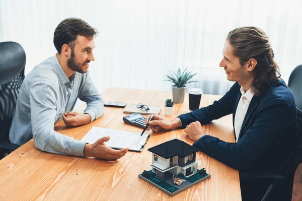 House model is displayed on wooden meeting table with in the blurred background of real estate agent and client discuss terms and conditions of house loan or rental lease contract. Entity