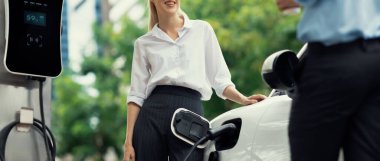 Progressive businessman and businesswoman with electric car parking and connected to public charging station before driving around city center. Eco friendly rechargeable car powered by clean energy.