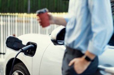 Şehirdeki halka açık elektrik şarj istasyonunda EV Odaklanma Otomobili 'nin çevreye duyarlı konsepti. Bulanık arka planda iş adamının arabasının kahvesini doldurmak için beklemesi..