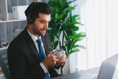 Çağrı merkezi operatörünün müdürü bilgisayarla uğraşırken masasında kahvesiyle oturuyor. Kulaklık takan ve siyah takım giyen bir ofis çalışanı müşteri desteği ya da tele pazarlama üzerinde çalışıyor. coşkulu