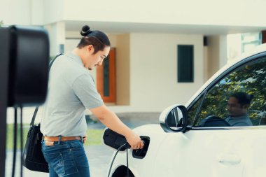 Progressive asian man install cable plug to his electric car with home charging station in the backyard. Concept use of electric vehicles in a progressive lifestyle contributes to clean environment.