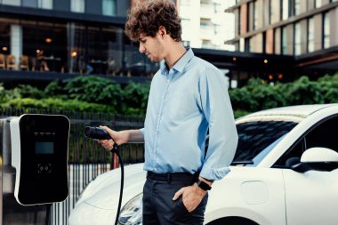 Progressive businessman insert charger plug from charging station to his electric vehicle with apartment condo building in background. Eco friendly rechargeable car powered by sustainable energy.