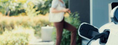 Focus EV charger plugged into EV car at home charging station with blurred background of progressive woman walking in background. Elective vehicle powered by clean energy for eco-friendly concept.