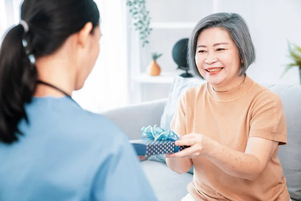 stock image A young caregiver hand over to her senior patient a blue gift box with blue ribbons at a contented living room.