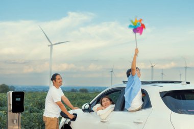 İlerici mutlu aile kavramı elektrikli araçla rüzgar çiftliğinde eğleniyor. Rüzgar türbini jeneratöründen gelen temiz yenilenebilir enerji ile şarj edilen elektrikli araç..