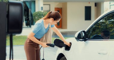 Progressive woman install cable plug to her electric car with home charging station. Concept of the use of electric vehicles in a progressive lifestyle contributes to clean environment.