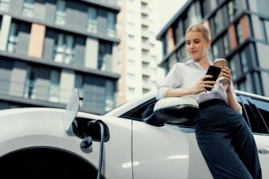 Focus businessman using phone, leaning on electric vehicle, holding coffee with blurred city residential condo buildings in background as progressive lifestyle by renewable and sustainable EV car.