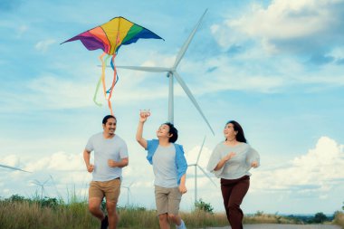 Progressive happy carefree family vacation concept. Young parents mother father and son run along and flying kite together on road with natural scenic on mountain and wind turbine background.