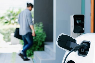 Focus electric car charging at home charging station with blurred progressive man walking in the background. Electric car using renewable clean for eco-friendly concept.