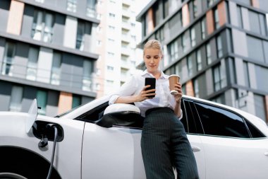 Focus businessman using phone, leaning on electric vehicle, holding coffee with blurred city residential condo buildings in background as progressive lifestyle by renewable and sustainable EV car.