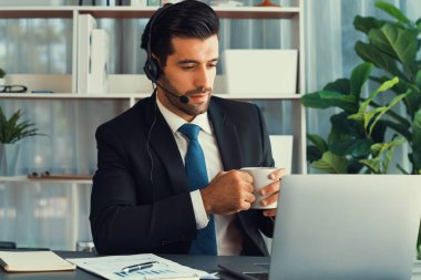 Çağrı merkezi operatörünün müdürü bilgisayarla uğraşırken masasında kahvesiyle oturuyor. Kulaklık takan ve siyah takım giyen bir ofis çalışanı müşteri desteği ya da tele pazarlama üzerinde çalışıyor. coşkulu
