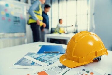 Concept of engineering equipments of protective helmet for competent engineer on piles of paper works. Blurred image of engineers and employer working together at the desk.