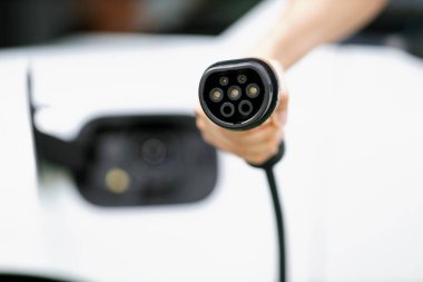 Progressive new alternative clean energy vehicle and electric charging device concept. Focus closeup hand holding and pointing EV charger plug at camear from electric charging station at camera