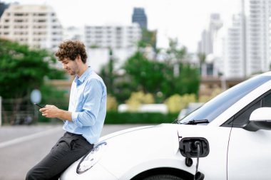 Progressive businessman talking on the phone, leaning on electric car recharging with public EV charging station, apartment condo residential building on the background as green city lifestyle.