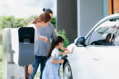 Progressive young parents and daughter with electric vehicle and home charging station. Green and clean energy from electric vehicles for healthy environment. Eco power from renewable source at home.